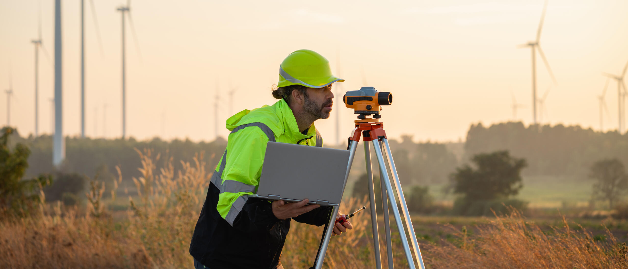 Field surveyor using a laptop and leveling equipment during an outdoor site survey.