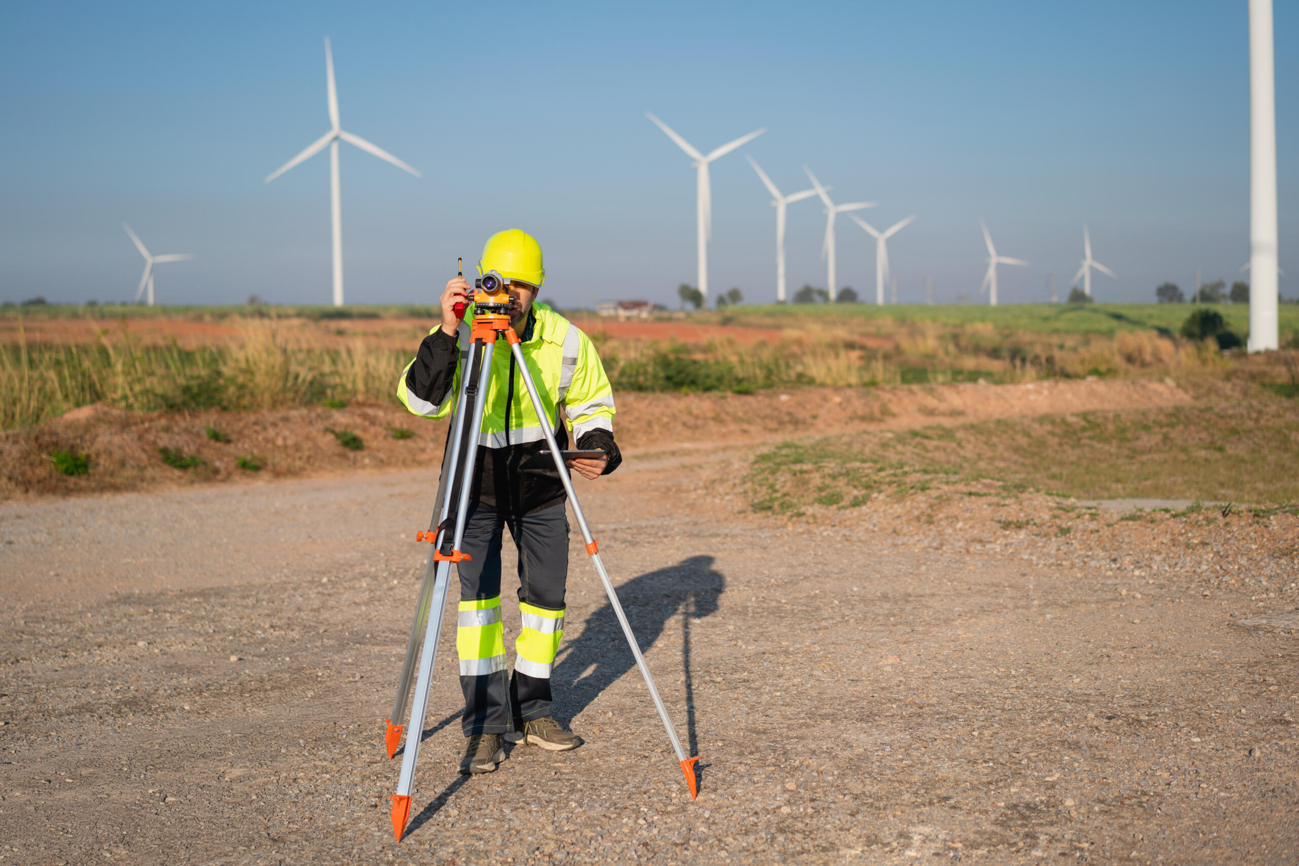 Surveyor using land surveying equipment in an open field near wind turbines.