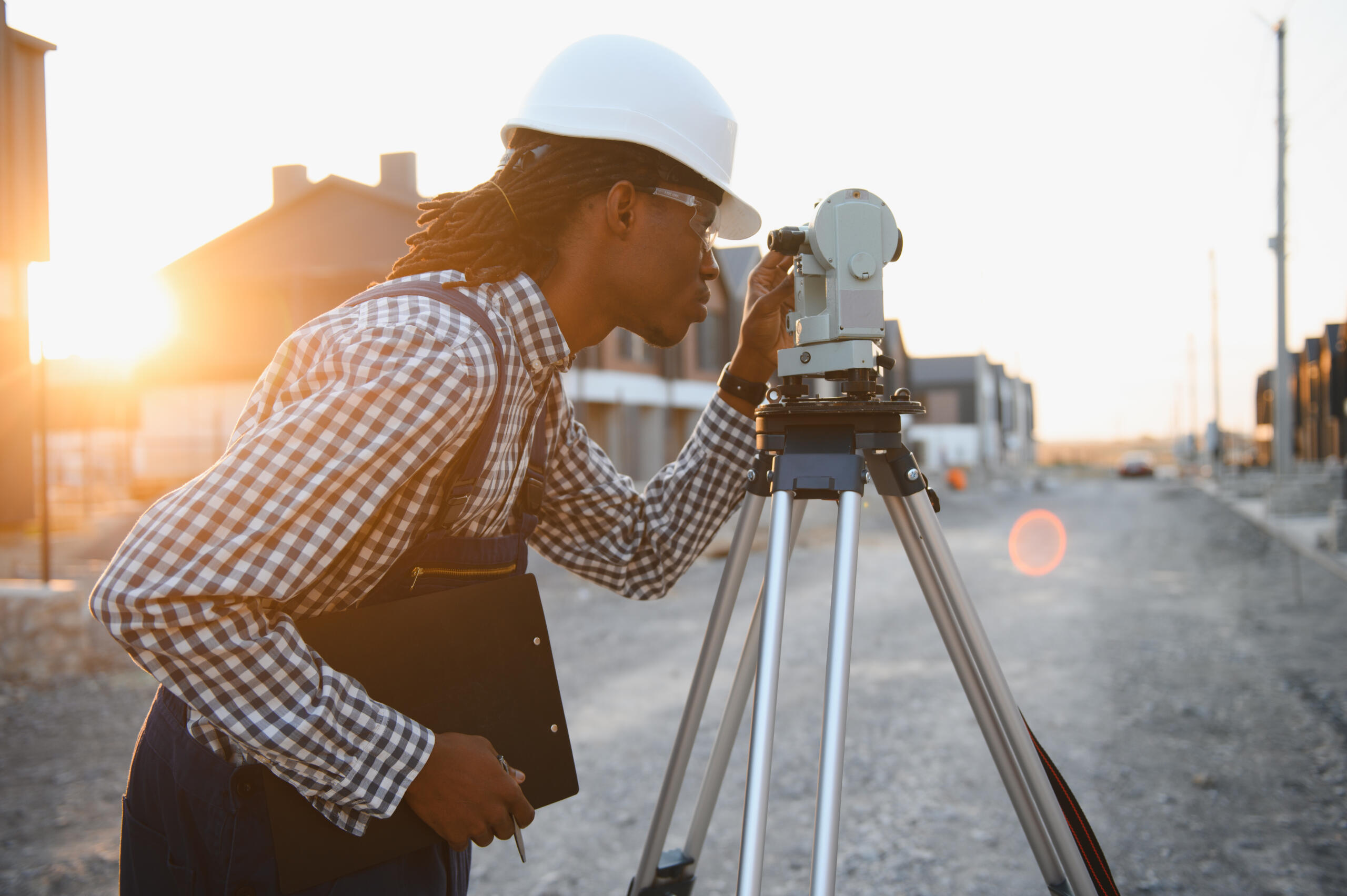 A surveyor in a white hard hat looks through a total station on a tripod at a residential construction site