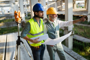 Two workers in hard hats holding blueprints stand next to a surveying instrument on a tripod within a concrete building structure, utilizing professional land survey services for construction projects to plan site layout.