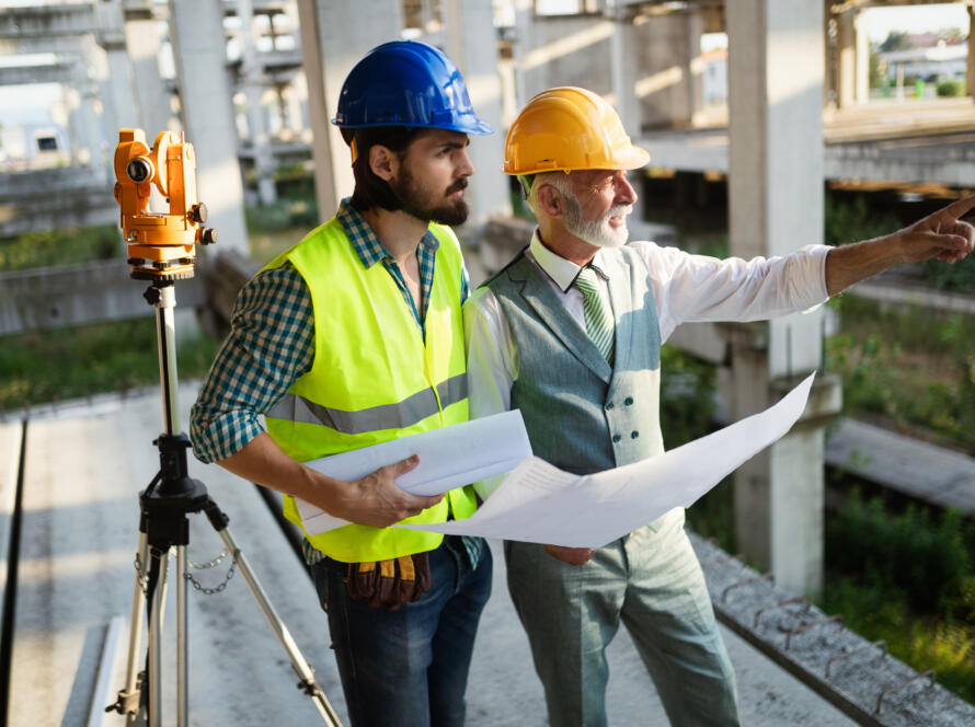 Two workers in hard hats holding blueprints stand next to a surveying instrument on a tripod within a concrete building structure, utilizing professional land survey services for construction projects to plan site layout.