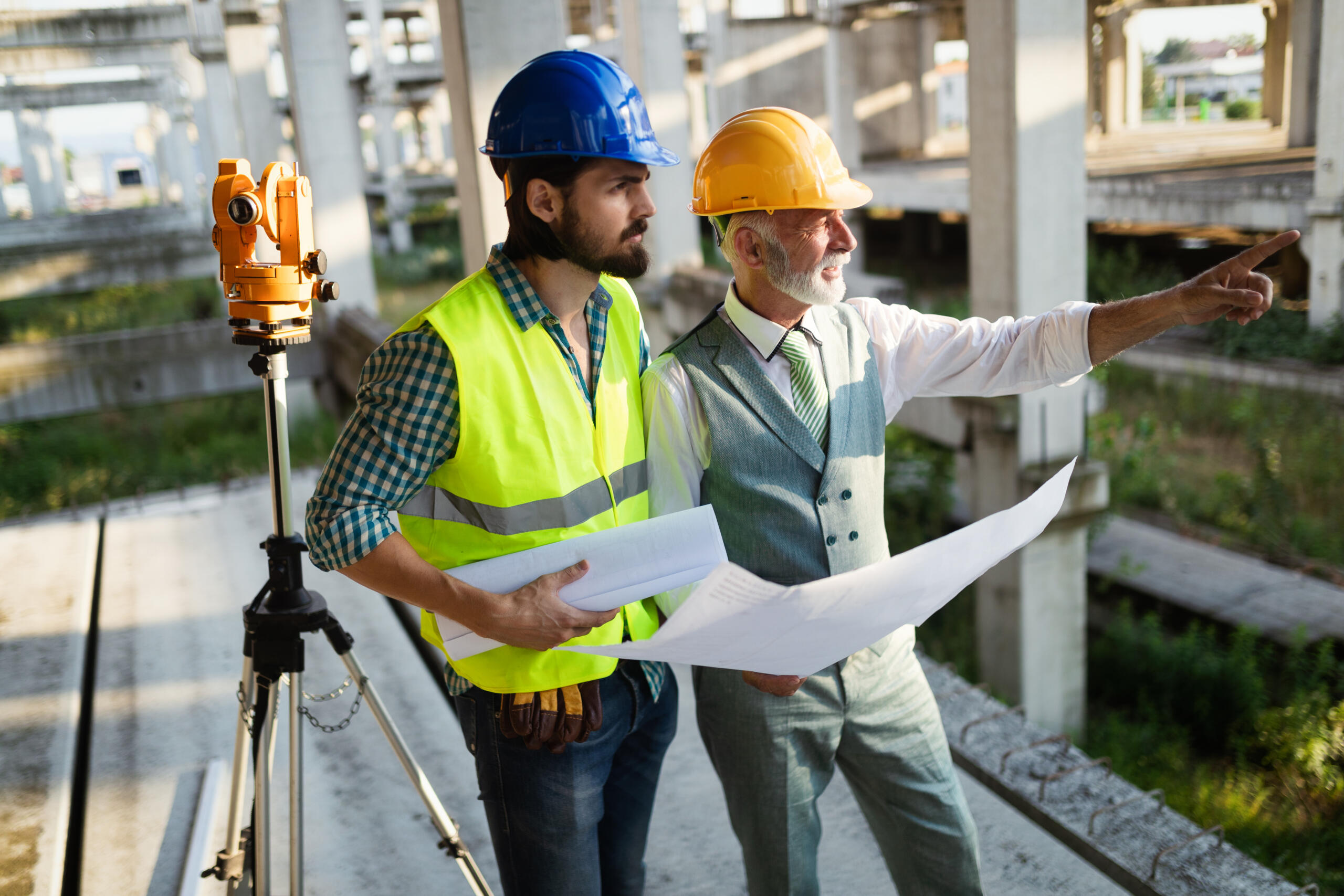 Two workers in hard hats holding blueprints stand next to a surveying instrument on a tripod within a concrete building structure, utilizing professional land survey services for construction projects to plan site layout.