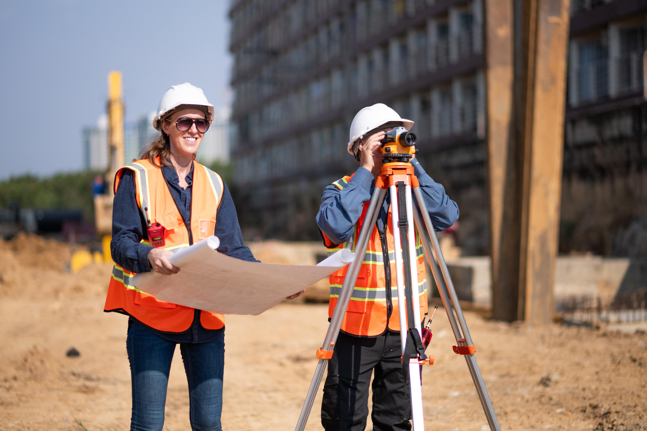 Two construction professionals in hard hats and orange vests work at a building site