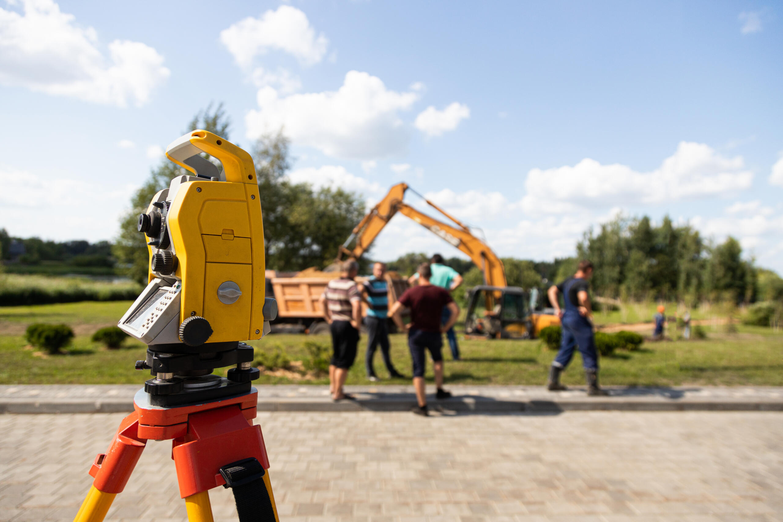 A close-up side view of a yellow total station on an orange tripod, utilizing construction staking services on a site where workers stand near a dump truck and excavator