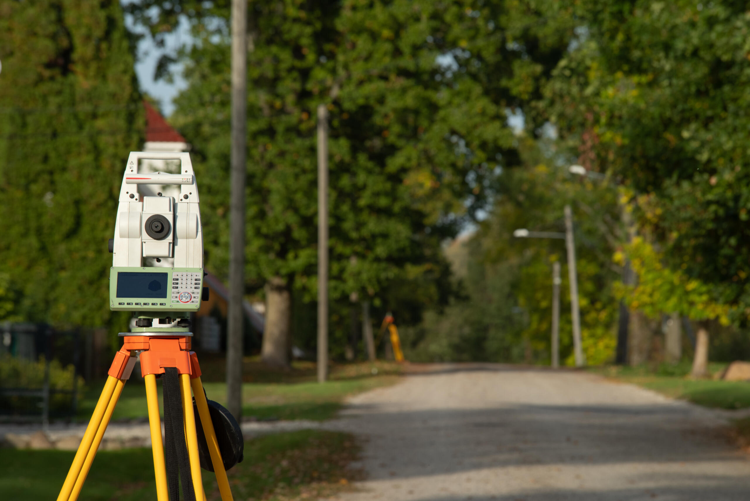 A white digital total station surveying instrument mounted on a yellow and orange tripod sits along the side of a rural road bordered by green trees