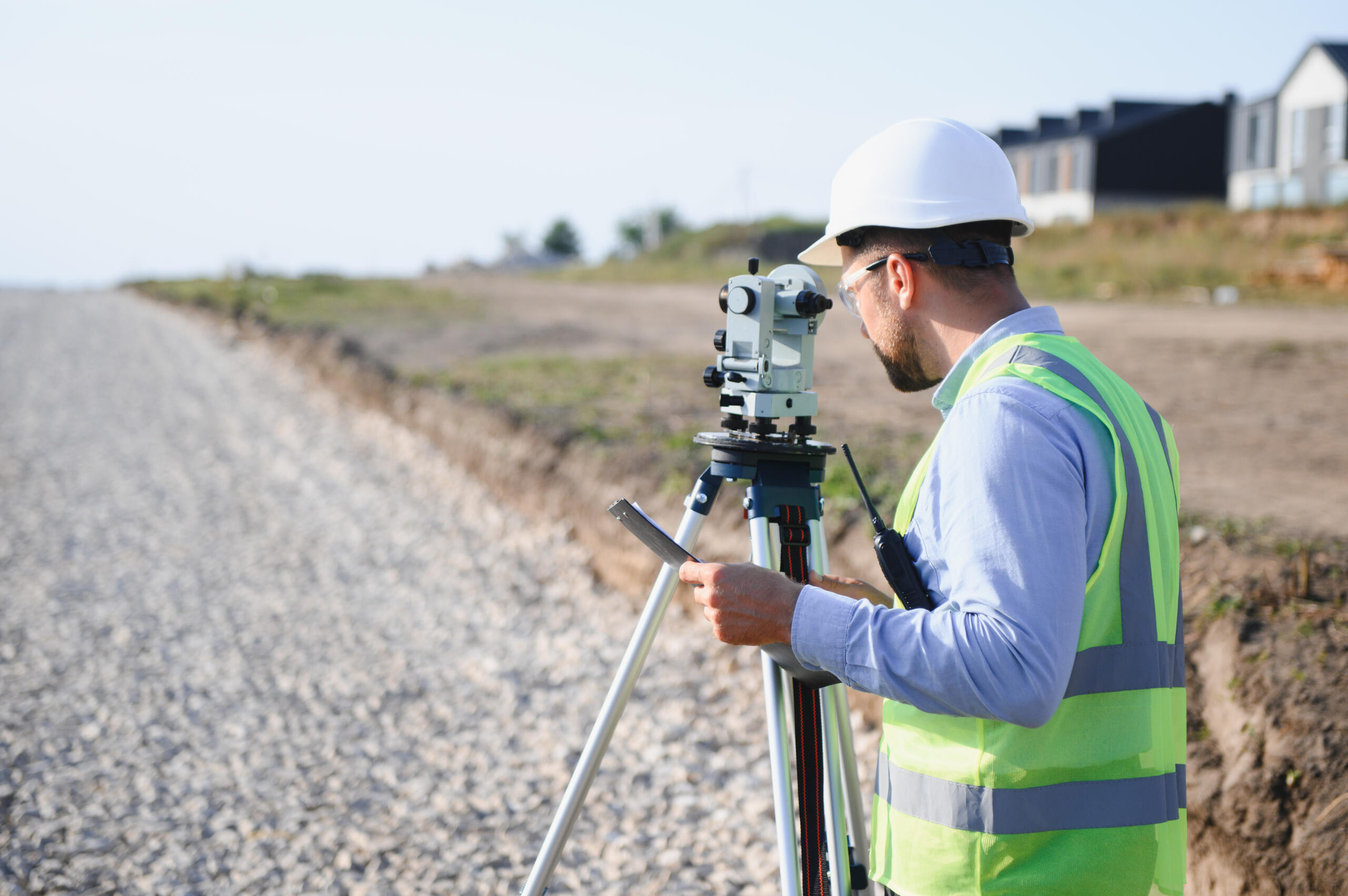 surveyor wearing a white hard hat and a high-visibility green vest looking through a total station level on a tripod