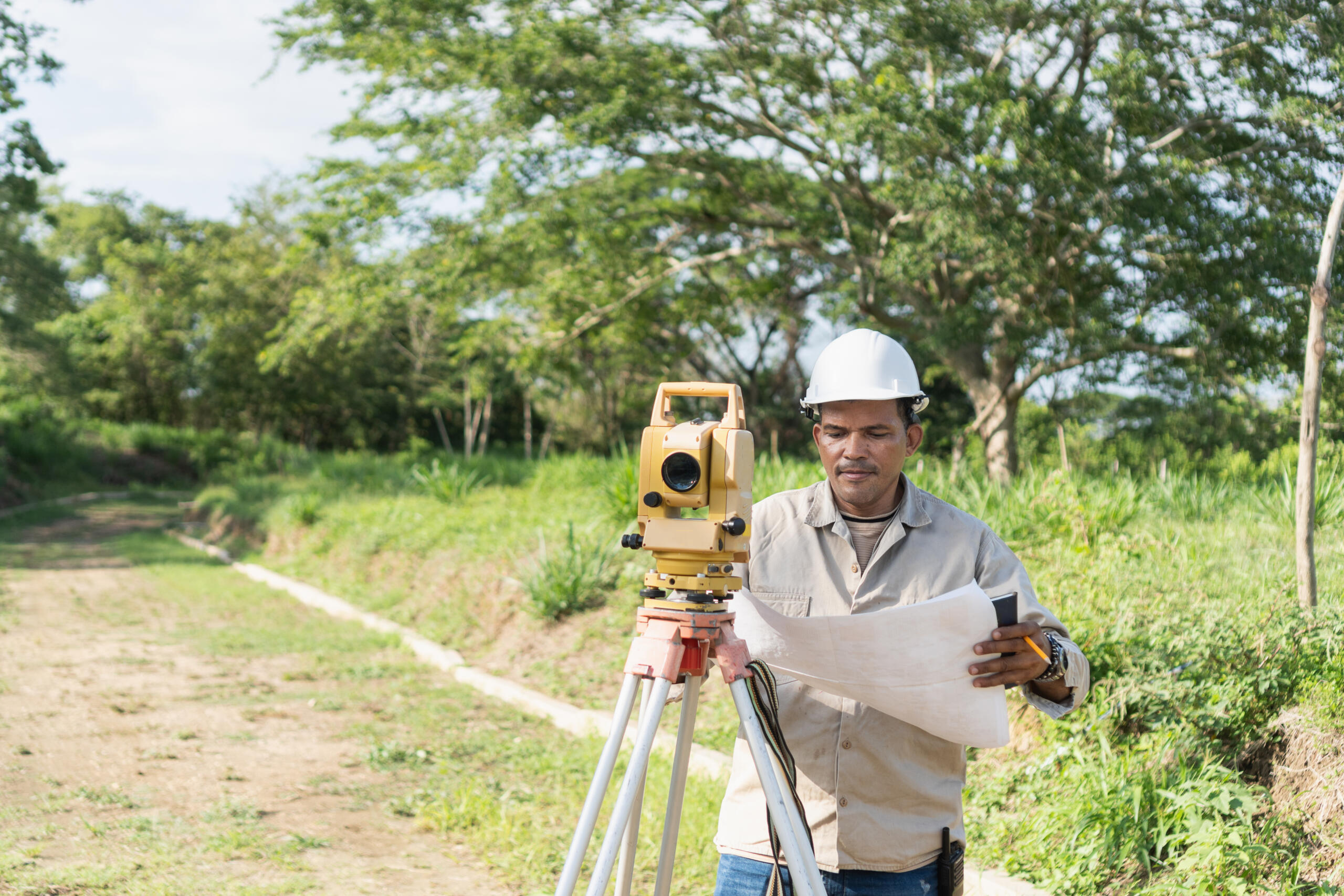 A surveyor in a hard hat reviews maps next to a yellow total station on a tripod in a green field, a standard procedure when you request professional land survey services South Texas projects require.