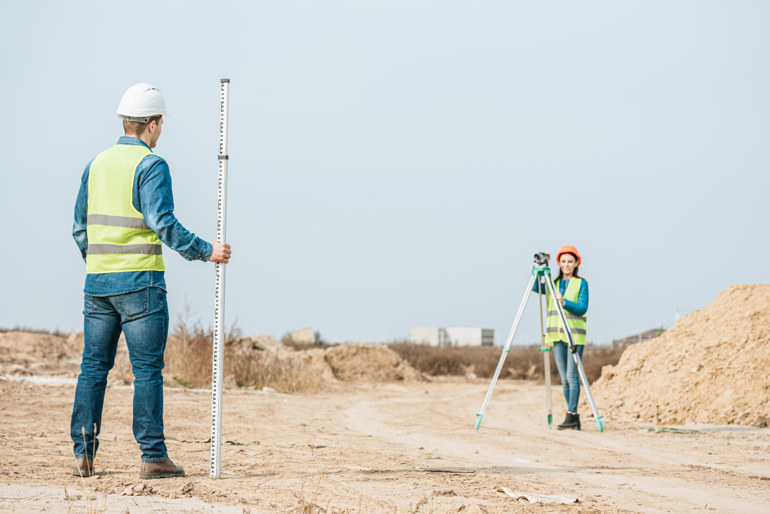 A male surveyor wearing a white hard hat holds a tall leveling rod in the foreground, while a female surveyor in an orange hard hat operates an optical level on a tripod in the background.