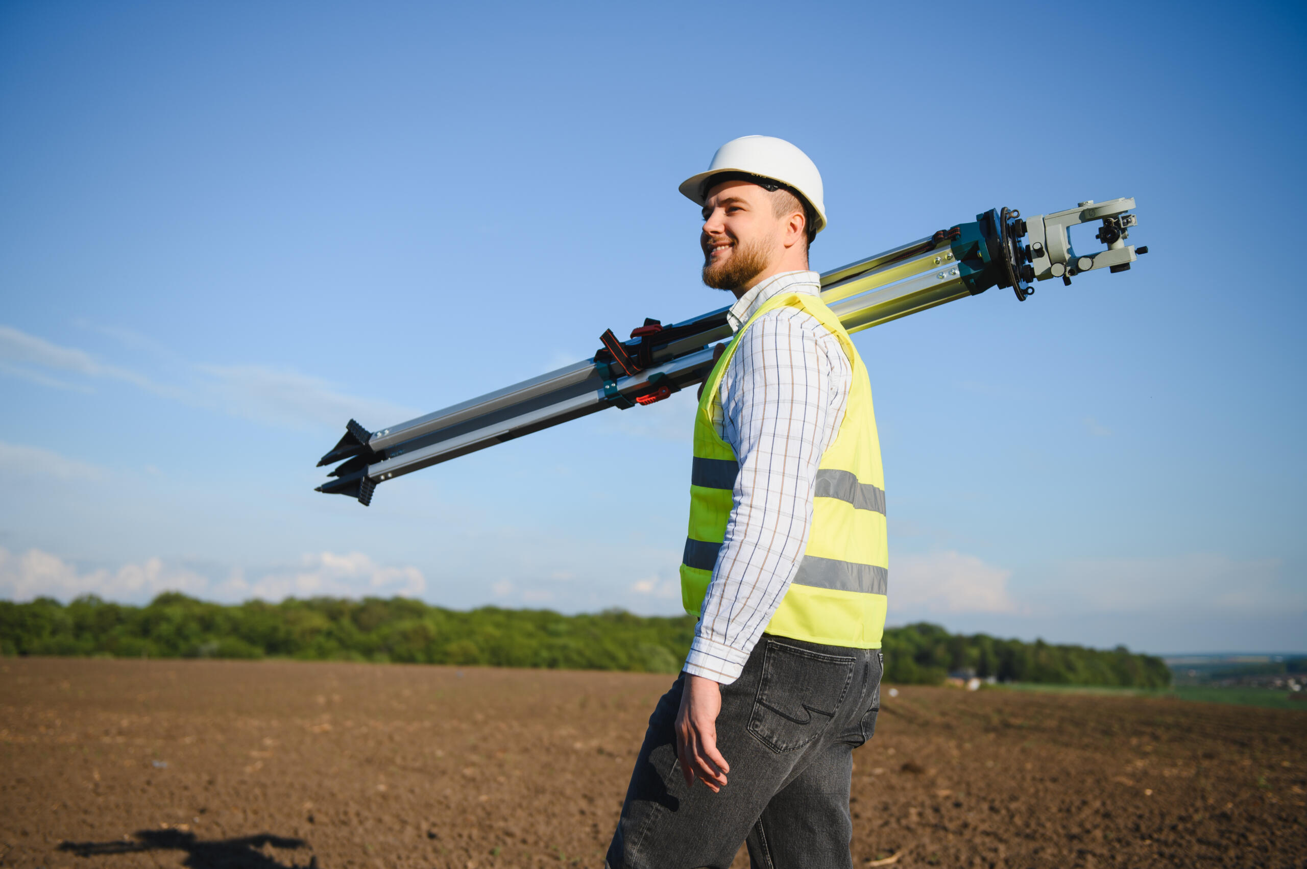 A smiling surveyor wearing a white hard hat and yellow safety vest walks through an open field while carrying a tripod over his shoulder, delivering professional land surveying services
